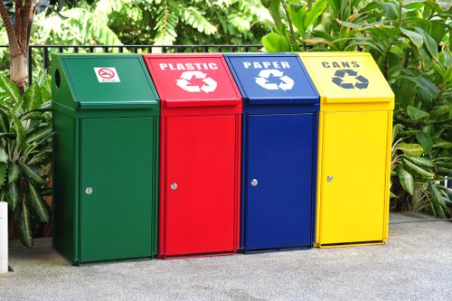 The image displays four colour-coded waste disposal bins positioned on a paved outdoor surface, set against a backdrop of green foliage including shrubs and small trees, suggesting a garden or residential setting near Gunnersbury. The first bin on the left is green and designated for general waste, featuring a white symbol with a red circle and cross indicating no littering. Next is a red bin marked for plastic waste, with a white recycling symbol and the word 'PLASTIC' printed above. The third bin is blue for paper recycling, also displaying a white recycling symbol and the label 'PAPER.' The fourth bin, yellow in colour, is designated for cans, marked with a white recycling symbol and the word 'CANS.' Each bin has a small, round, silver keyhole lock on the front and a slightly sloped lid. The bins are made of durable plastic with a textured finish, and their arrangement suggests a community or commercial rubbish collection setup, which is supported by the services offered by Commercial Waste Gunnersbury. Overall, the scene emphasizes proper waste segregation in line with local recycling practices that a waste management specialist would support in Gunnersbury and surrounding areas.