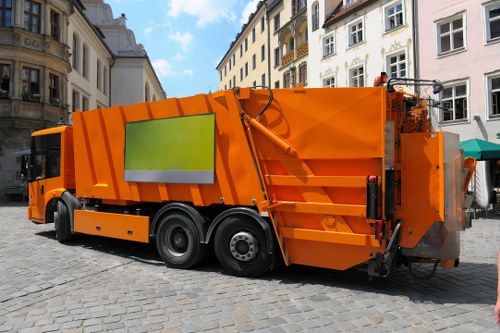 A large orange commercial waste collection vehicle parked on a cobbled street in an urban area, with multi-storey white and beige buildings in the background. The vehicle features a reflective yellow-green safety strip on its side, with a smooth, metallic finish and visible on-board compaction mechanisms at the rear. The street appears to be in a city setting, possibly in or near Gunnersbury, with the vehicle positioned centrally on the paved surface. Surrounding structures include windows and balconies typical of residential or mixed-use buildings, and the scene is lit by natural daylight, creating clear shadows on the ground. This image visually supports rubbish removal services provided by Commercial Waste Gunnersbury, illustrating a street collection operation in a London-based urban environment.