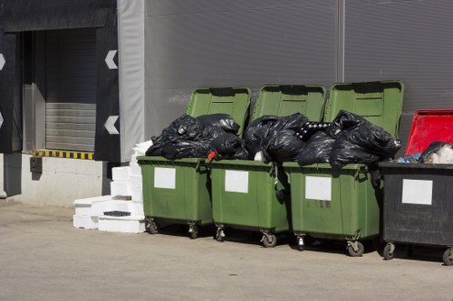 Four large wheelie bins positioned on a concrete surface in an urban or industrial setting, with three of the bins in green and one in black. The green bins are filled with black rubbish bags overflowing from the open lids, indicating they are used for general waste collection. The black bin has a closed lid and contains additional waste. Behind the bins, there are metallic grey roller shutters and a wall with grey cladding, suggesting the location could be a commercial or storage premises. To the left, a stack of white bricks or concrete blocks is visible on the ground, partially obscured by the bins. The scene is outdoors, with natural lighting and minimal environmental context, conducive to rubbish removal services operating in the Gunnersbury area or nearby London districts. The presence of waste bins ready for collection and the clean, organized arrangement reflect typical commercial waste management activities in the local area, as handled by companies such as Commercial Waste Gunnersbury.