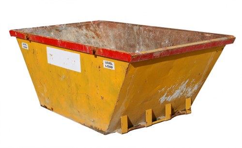 A large, industrial-style skip with a rectangular shape and sloped sides, placed outdoors on a paved surface. The skip is painted yellow with a red-painted rim at the top, and it shows signs of dirt and rust along the exterior surface, indicating frequent use in waste collection. The interior of the skip is visible, revealing a rusty, metallic surface with a rough texture. Mounted on the front is a white label area, currently blank, and a black-and-white sign indicating maximum load capacity. The skip is positioned in an open area, possibly near a commercial or industrial site, with no other objects or background scenery visible in the image. The lighting suggests daylight, with natural illumination highlighting the weathered finish of the skip, which relates to waste removal services provided by Commercial Waste Gunnersbury, serving local businesses in the area near W12 postcodes.