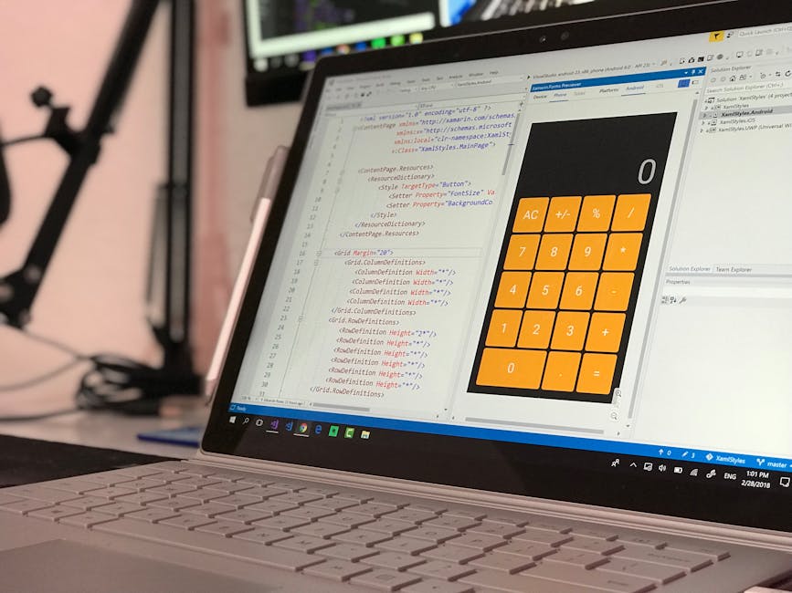A close-up view of a laptop computer on a desk, displaying a software development environment with lines of HTML code in a text editor on the left side of the screen. The right side shows a mobile app